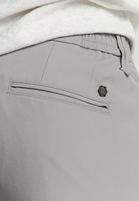 Grijze broek van gladde stof heeft een enkele achterzak met een rechthoekige opening en een zwart-gouden hexagonaal logo detail.