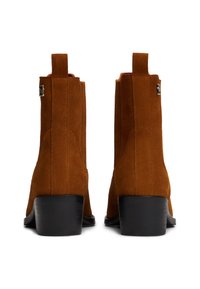 Bottines en daim marron avec un talon carré, dotées d'anneaux de tirage latéraux, de détails cousus et d'un accent argenté à l'extérieur.