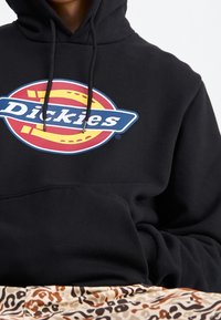 Osoba v černé mikině s barevným logem Dickies a béžových vzorovaných kalhotách, s částí rukou viditelnou v kapse mikiny.