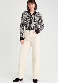 Chemise à motifs noir et blanc avec des manches longues et un col noir, assortie à un pantalon large crème et des bottes à talons noires.