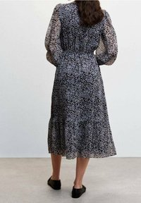 Zwart bloemenjurk met doorschijnende lange mouwen, een geplooid middel en een gelaagde rok. Bevat kleine witte en lichtpaarse bloemen. Platte zwarte schoenen.