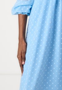 Vaaleansininen mekko, joka on valmistettu läpikuultavasta kankaasta, jossa on hienot valkoiset pilkut. Mekko sisältää puffipaidan hihat ja löysän istuvuuden.