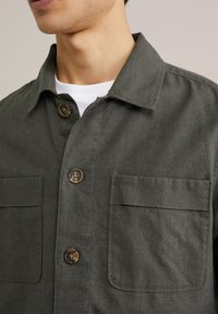 Donkergroene shirtjack vervaardigd uit getextureerde stof, met een puntige kraag, twee borstzakken en bruine knopen.