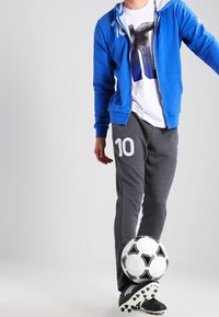 Sweat à capuche zippé bleu, t-shirt blanc avec graphisme, pantalon de survêtement gris avec impression "10", et crampons de football noirs. Tenant un ballon de football noir et blanc.