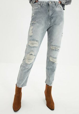 Vervaagde grijze gescheurde jeans gedragen met bruine suède enkellaarzen, handen in de zakken gestoken, staand tegen een effen witte achtergrond.