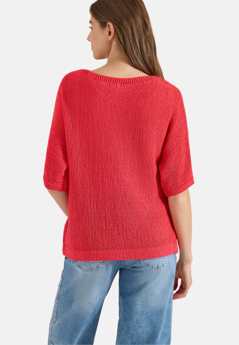 Femme aux longs cheveux bruns portant un haut en maille rouge ample et un jean bleu clair, vue de dos sur un fond uni.