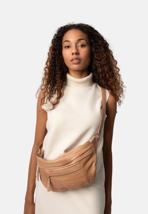Borsa a tracolla in pelle marrone chiaro con due tasche con zip, forma curva e tracolla regolabile, indossata sopra un maglione a collo alto color crema chiaro.