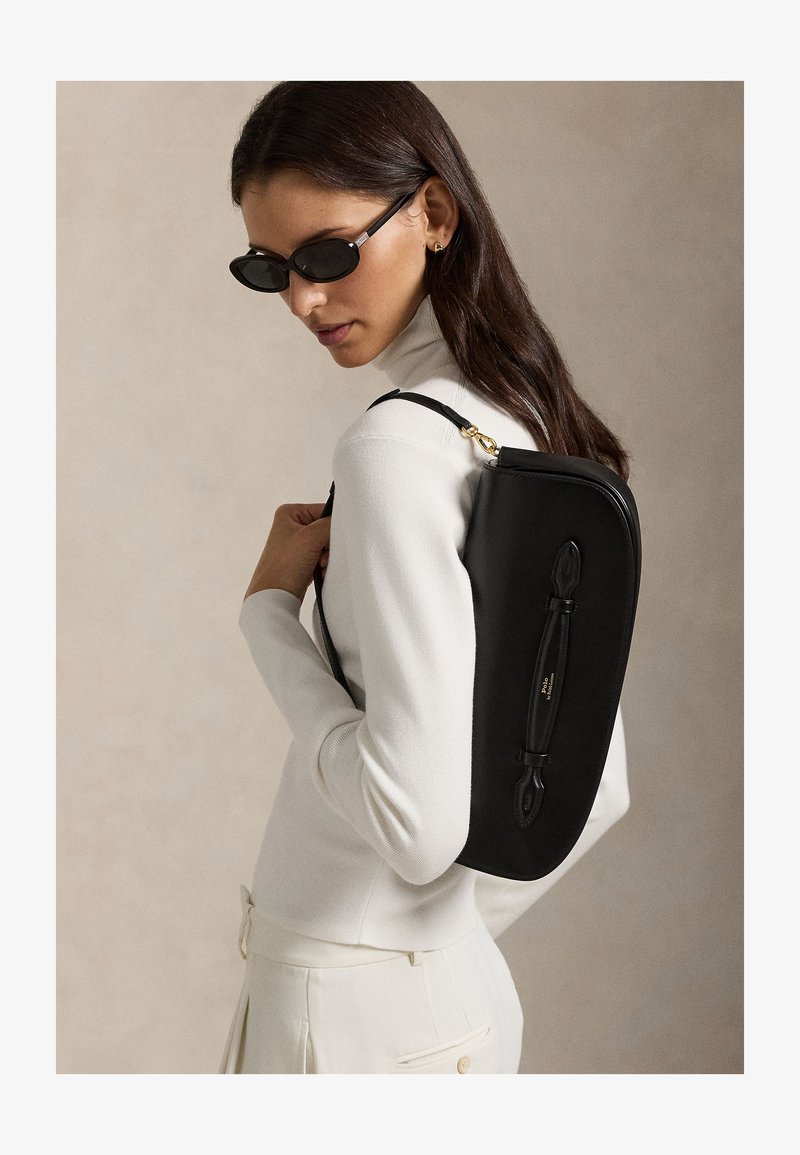 Woman wearing black sunglasses and white turtleneck carries a sleek black shoulder bag against a neutral background.