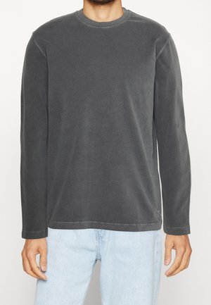 Man draagt een antracietgrijs shirt met lange mouwen en ronde hals, gecombineerd met lichtblauwe spijkerbroek, staand tegen een effen achtergrond.