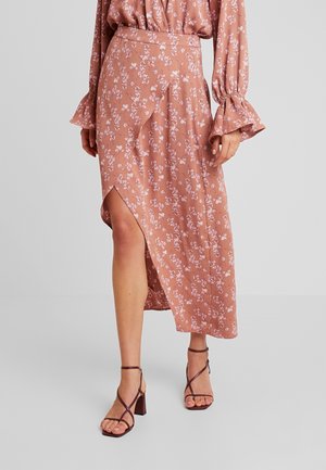 Femme portant une jupe portefeuille rose à fleurs avec une fente et un chemisier assorti avec des poignets volantés, associée à des sandales à talons hauts marron foncé à brides.