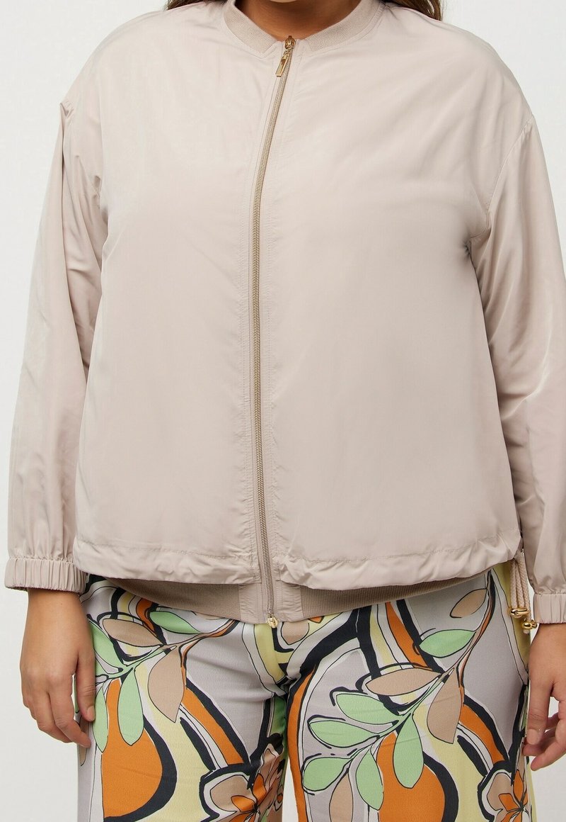 Haut du corps portant une veste zippée beige associée à un pantalon coloré avec un motif abstrait de feuilles et géométrique.