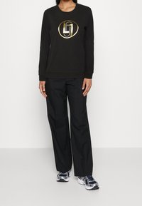 Sudadera negra con logo circular dorado y detalles texturizados, combinada con pantalones negros y zapatillas deportivas azules y blancas.