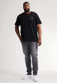 Camiseta negra con un logo dorado en el lado izquierdo del pecho, combinada con jeans gris desgastados y zapatillas deportivas blancas. Diseño sencillo y casual con un corte relajado.