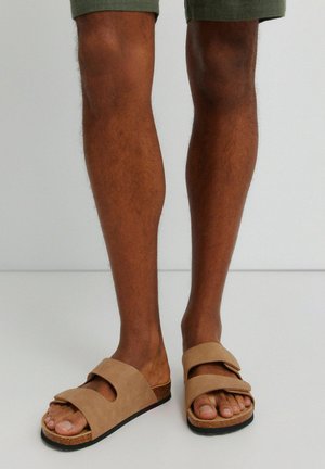 Legs wearing olive green shorts and tan open-toe sandals with two adjustable straps and cork soles on a light background.