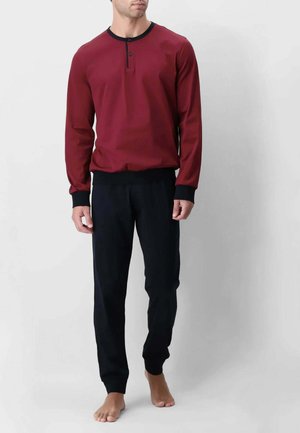 Uomo che indossa una maglia henley bordeaux a maniche lunghe con polsini neri e pantaloni neri, in piedi a piedi nudi su uno sfondo semplice.