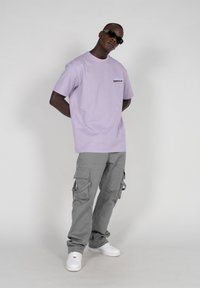Tricou din bumbac lavandă cu un mic logo negru, asortat cu pantaloni cargo gri deschis, cu multiple buzunare, și adidași albi.