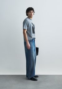 Gray t-shirt with a front graphic, loose blue denim jeans with a faded look, and black slip-on shoes, holding a black folder.