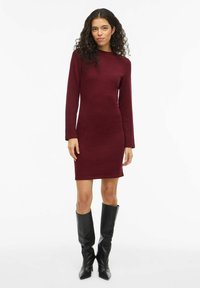 Vestito bordeaux in maglia con maniche lunghe e scollo rotondo. Silhouette aderente, abbinato a stivali neri al ginocchio. Tessuto liscio.