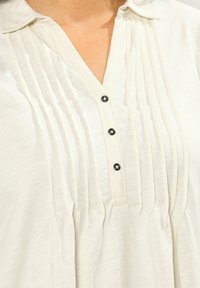 Krämfarbene Bluse mit Kragen, vertikalen Falten und drei kleinen schwarzen Knöpfen auf der Vorderseite, getragen auf nackter Haut.