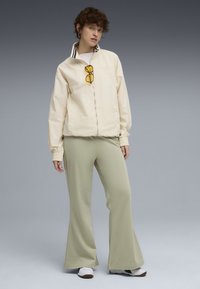 Lichtbeige zip-up jack met een gestreepte kraag, gecombineerd met lichtgroene flared broeken. Het model draagt gele zonnebril en witte sneakers.