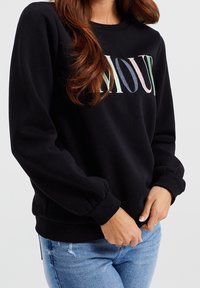 Svart sweatshirts med långa ärmar, ribbade ärmslut och rund halsringning. Har "AMOUR" tryckt i flerfärgade bokstäver på framsidan.