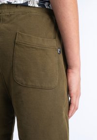 Pantalons vert olive avec un tissu texturé. Présente une poche arrière bien visible avec des bords arrondis et une petite étiquette de marque sur la poche.