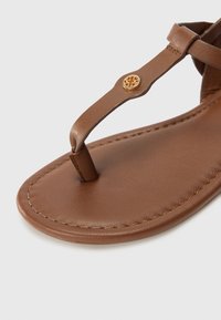 Brun läder sandal med tånoursdesign. Har en cirkulär gyllene detalj och kontrasterande sömmar längs kanten. Slät textur.
