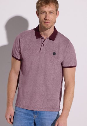 Mann trägt ein hellviolettes Polohemd mit dunkelviolettem Kragen und Ärmelsäumen, kombiniert mit hellblauen Jeans, steht vor einem einfarbigen Hintergrund.