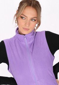 Gilet in fleece viola con colletto alto, zip completa e una texture morbida. Indossato sopra un top nero a maniche lunghe, dal design aderente.