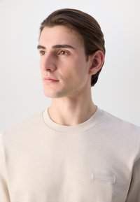 Felpa leggera beige chiaro con scollo rotondo e una texture sottile. Presenta una piccola etichetta con logo sulla parte anteriore, design minimalista.