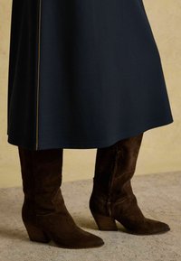 Merineel kleit suju tekstuuriga ja külgõmbluste detailiga, koos pruunide suede kuni põlveni saapadega, millel on kitsendatud varvas ja kihiline konts.