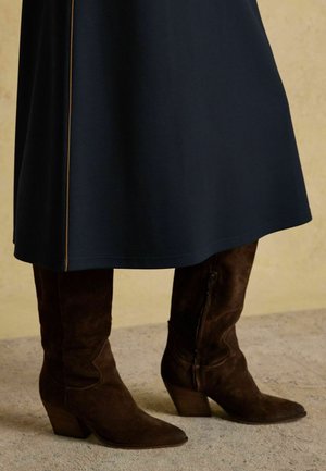 Navyblaues Kleid mit glatter Textur und seitlichem Nahtdetail, kombiniert mit braunen Wildleder, knielangen Stiefeln mit spitzer Zehenpartie und gestapeltem Absatz.