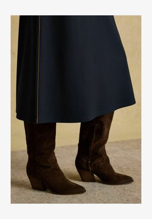 Navyblaues Kleid mit glatter Textur und seitlichem Nahtdetail, kombiniert mit braunen Wildleder, knielangen Stiefeln mit spitzer Zehenpartie und gestapeltem Absatz.