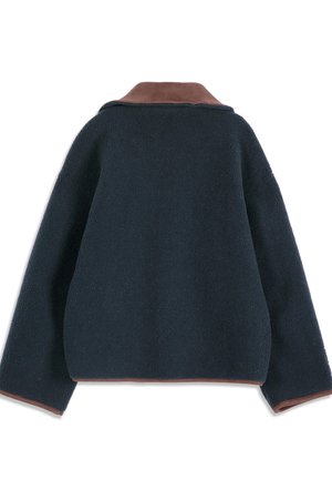 Jeune femme debout portant une veste oversize bleu marine foncé, un pull violet à motifs, un pantalon large en velours côtelé gris et des bottes camel avec des semelles noires.