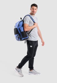 Mochila de tejido azul y negro, con detalles de malla, múltiples compartimentos y correas ajustables. Hombre con camiseta gris y pantalones negros.