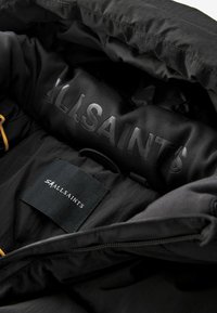 Unutrašnjost crne izolirane jakne s glatkom teksturom. Sadrži urezanu oznaku "ALLSAINTS" i etiketu za njegu tkanine. Zipper s tekstilnim rubovima.