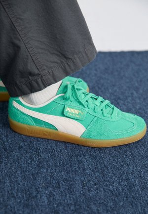Pieds portant des baskets Puma Palermo vertes avec des chaussettes blanches et un pantalon foncé sur un sol recouvert de moquette bleue.