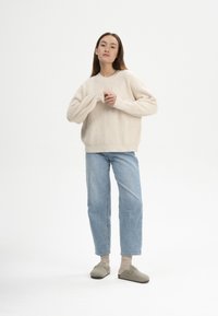 Helle beige Strickpullover mit weiten Ärmeln, kombiniert mit hellblauen Jeans im geraden Schnitt und grauen Clogs. Modell steht vor einem weißen Hintergrund.