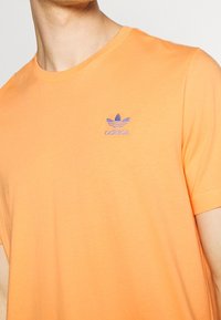 Oranžové bavlněné tričko s kulatým výstřihem a krátkými rukávy; má na levé straně hrudi vyšitou fialovou značku Adidas.