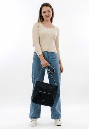 Sac en cuir noir avec une poche zippée à l'avant, surface texturée et doubles poignées, tenu par une personne en pull léger et jeans.