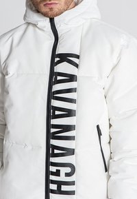 Bijela pufasta jakna s crnim zatvaračem i istaknutim logom "KAVANAGH" sprijeda. Ima kapuljaču i bočne džepove, glatke teksture.