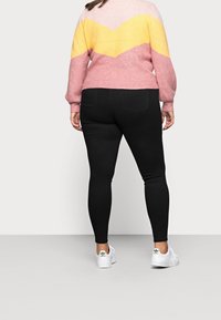 Person som bär en rosa och gul zigzagtröja, svarta tjejer jeans och vita Adidas sneakers, står vänd mot en enkel ljusgrå bakgrund.