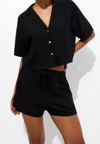 Chemise noire en lin à manches courtes avec col et boutons sur le devant, associée à un short noir assorti à cordon de serrage avec poches latérales.