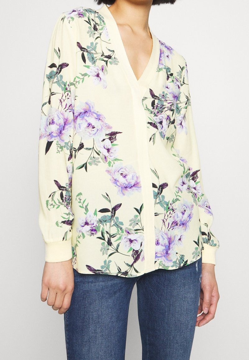 Blusa floreale gialla con design viola e verde, scollatura a v, maniche lunghe a sbuffo e polsini elastici. Abbinata a jeans di denim.
