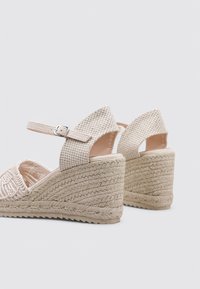Sandalias de cuña beige con parte superior de tela trenzada, correa en el tobillo con hebilla y un detalle de cuerda texturizada en el tacón de cuña.