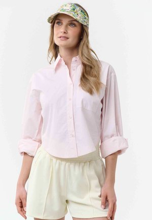 Mujer con cabello largo y rubio, vistiendo una camisa de botones rosa claro, pantalones cortos crema con cintura elástica y una visera colorida con estampado.