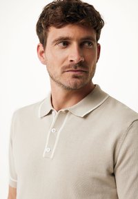 Béžové bavlněné polo tričko s klasickým límečkem, zapínáním na tři knoflíky a kontrastním bílým lemem na límci a rukávech.
