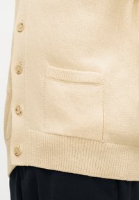 Μπεζ πλεκτή ζακέτα με κουμπιά μπροστά, μια μικρή τσέπη στο πλάι και ριμπ τελείωμα, φορεμένη πάνω από σκουρόχρωμο παντελόνι.