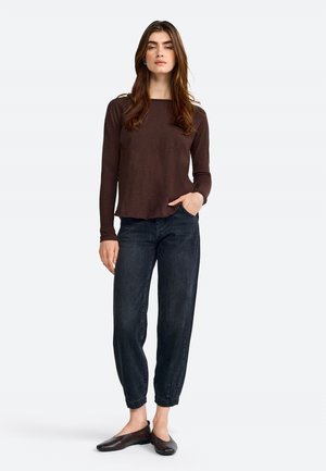 Top marrone a maniche lunghe con orlo arrotondato, abbinato a pantaloni in denim neri aderenti e ballerine scure. Il modello è in piedi su uno sfondo chiaro.