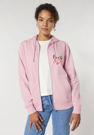 Veste hoodie zippée rose avec une poche frontale, ornée d'un graphique décoratif 'love' en noir et rouge. Portée sur un t-shirt blanc et un jean bleu.